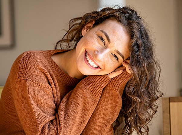 Woman smiling warmly in brown sweater, indoor setting with neutral background.