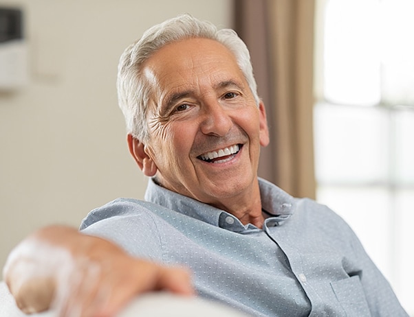Smiling older man in light blue shirt gestures welcomingly indoors, dental office setting.