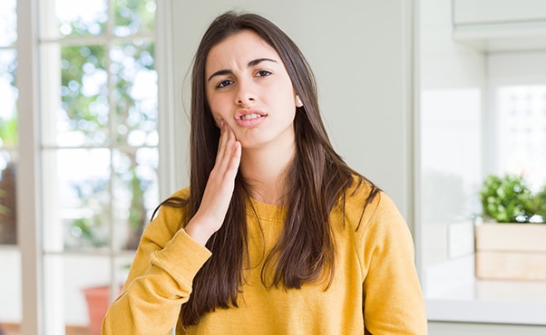Woman in yellow shirt touching face thoughtfully, indoors with bright natural light.