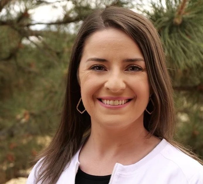 Professional female dental staff member smiling outdoors with evergreen trees in background.