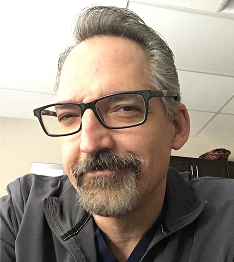 Professional man with glasses and beard in dental office setting, wearing dark shirt.