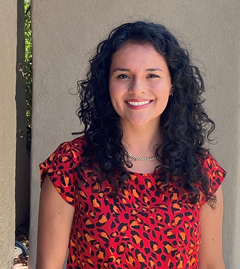 Woman with long dark curly hair smiling at camera wearing red patterned top against neutral background.