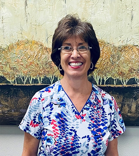 Female dental professional smiling at camera wearing colorful patterned scrubs in dental office setting.