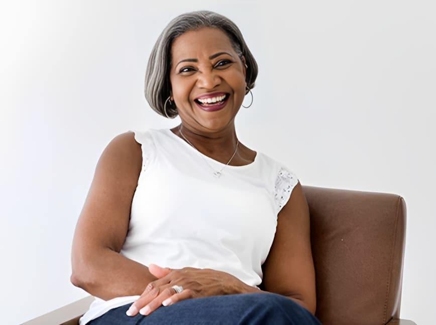 Woman smiling at camera seated in brown chair wearing white sleeveless top